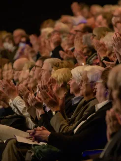 The audience at the DRK gala at Oper Köln sits in rows side by side, applauding enthusiastically. Many spectators are seen in profile with raised hands and focused gazes directed toward the stage. The scene is illuminated by warm stage lighting, creating a festive and appreciative atmosphere.