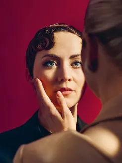 Close-up against a red background with warm, cinematic lighting. At the center stands an androgynous-looking person with short, auburn hair styled in soft finger waves in a dark jacket. Their blue eyes gaze calmly toward the viewer, serious expression appearing thoughtful. In the right foreground, a blonde person with a braid and a light-colored blazer is visible out of focus. Their hand rests gently on the central figure’s cheek. The intimate closeness evokes The Knight of the Rose by Richard Strauss.