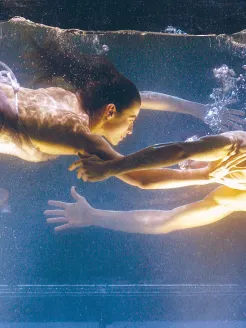 Underwater scene from Dido and Aeneas: two performers float facing each other in a water tank, their bodies gently suspended, arms reaching toward one another as air bubbles rise between them and warm, golden light fills the scene.