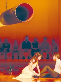 Scene from The Coronation of Poppea by Claudio Monteverdi: Poppea kneels in a light-colored dress, leaning intensely toward Nero, who sits opposite her and gazes at her attentively. Warm orange lighting creates a sensual and tension-filled atmosphere. In the background, a row of figures watches the scene unfold. A large cylindrical stage element emphasizes the production’s minimalist, modern aesthetic.