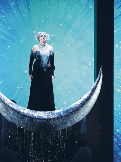 Scene from The Magic Flute by Wolfgang Amadeus Mozart: The Queen of the Night stands on a silver crescent moon in a sparkling dark gown, set against a radiant, star-like backdrop; her presence appears majestic and powerful.