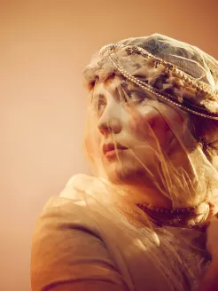 Close-up of a young bride in warm, cinematic lighting. She wears a transparent veil adorned with pearls, softly framing her face. Her gaze is directed slightly upward to the left, as if looking toward an uncertain future. Her expression combines longing and determination. Warm tones, delicate fabric textures, and soft light create an intimate, poetic atmosphere. Inspired by Marie from Smetana’s opera The Bartered Bride.