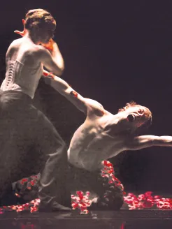 In a darkly staged scene, warm, focused lighting highlights the bodies of two dancers. In the foreground, a shirtless dancer kneels with his torso arched far backward, arms outstretched, head thrown back, and mouth open—an expression of extreme tension. Beside him stands a female dancer wearing a white corset and dark trousers, grasping his arm. Red flower petals cover the floor. The scene feels highly expressive, sculptural, and charged with both conflict and devotion.