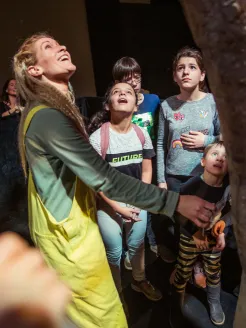 Eine Gruppe von Kindern und eine erwachsene Begleitperson stehen während einer Familienführung im Opernhaus vor einem großen Objekt. Die Teilnehmerinnen und Teilnehmer blicken aufmerksam nach oben und wirken neugierig und fasziniert. Die Begleitperson zeigt auf etwas außerhalb des Bildausschnitts. Die Szene ist warm ausgeleuchtet und vermittelt eine Atmosphäre des gemeinsamen Entdeckens und Staunens.