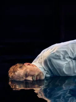 Nahaufnahme eines Mannes mittleren Alters vor schwarzem Hintergrund in warmer, cineastischer Lichtstimmung. Mit kurzem, zerzaustem braunem Haar liegt er auf einem wasserbedeckten Boden und blickt den Betrachter an. Er trägt ein weißes Hemd; Gesicht und Oberkörper spiegeln sich in der dunklen Wasserfläche. Sein erschöpfter, introvertierter Ausdruck vermittelt Rückblick, Sehnsucht und innere Zerrissenheit. Die Szene erinnert an Hoffmann aus Jacques Offenbachs Hoffmanns Erzählungen.
