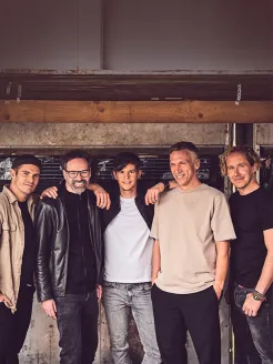 The band Kasalla is shown as a group of five men standing closely together, some with their arms around each other’s shoulders. They wear casual clothing such as T-shirts, shirts, and a leather jacket, and look relaxed into the camera. In the background, a rustic wall with wood and metal elements creates a warm, informal atmosphere.