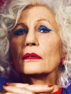 Close-up of an elderly woman in a warm, cinematic lighting. She looks into the camera with a determined and defiant expression. Silver-gray, curly hair frames her face. Dramatic stage makeup, featuring royal blue eyeshadow and bold red lipstick, creates striking accents. Fine lines and wrinkles add character and expressiveness. A slightly out-of-focus hand in the foreground adds depth to the composition. The high-contrast, theatrical staging evokes Rachel from Hans Werner Henze’s La Piccola Cubana.