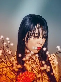 Close-up of a young Asian woman in warm, cinematic lighting. With long black hair and straight bangs, she gazes thoughtfully to the side in half profile. Her face is softly illuminated, while her red lips provide a striking accent. She wears a light-colored garment. Delicate branches with white blossoms frame her face, and a red flower adds contrast. The blurred background enhances the quiet, melancholic, and poetic mood. Inspired by Cio-Cio-San from Puccini’s Madama Butterfly.