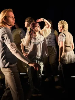 Mehrere Jugendliche und junge Erwachsene bewegen sich während einer Performance von Oper.Stadt.Schule über eine Bühne. Im Mittelpunkt steht eine Person in heller Kleidung, die eine ausdrucksstarke Pose einnimmt und von einem Lichtstrahl beleuchtet wird. Die anderen Teilnehmenden sind in Bewegung und umgeben die zentrale Figur. Die Szene ist kontrastreich ausgeleuchtet und vermittelt Dynamik, Kreativität und gemeinsames künstlerisches Arbeiten.