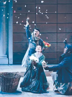 Scene from My Fair Lady by Frederick Loewe: On a Covent Garden flower market street set, three performers in period costume are surrounded by falling petals. An older man in coat, scarf, and cap tosses flowers into the air. In the foreground, Eliza Doolittle kneels in a long dress, holding white roses and looking up joyfully. Beside her, a young man in a dark coat and cap kneels with flowers in hand, gazing at her. The scene conveys the lively, festive atmosphere of a bustling flower market.