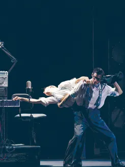 Several dancers appear in a stylized stage scene from the choreography Notte Morricone: At the center, a male dancer wearing glasses, a shirt, and suspenders carries a female dancer across his shoulders, while she arches her upper body backward and extends one arm. The scene is defined by props such as a microphone, a chair, and technical equipment, creating an atmosphere between stage and studio.
