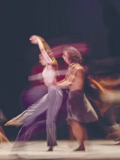 Several dancers in flowing motion on a stage; at the center, a duo dynamically spinning and lifting, with deliberately used motion blur, from the choreography Reversible by Annabelle Lopez Ochoa.