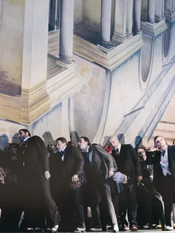 Scene from Rigoletto by Giuseppe Verdi: A group of elegantly dressed men in dark suits and tailcoats stand closely packed in front of a neoclassical architectural backdrop with columns and balustrades; their bodies lean forward, tense, as if reacting to something together.