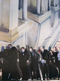 Szene aus Rigoletto von Giuseppe Verdi: Eine Gruppe elegant gekleideter Männer in dunklen Anzügen und Fräcken steht dicht gedrängt vor einer klassizistischen Architekturkulisse mit Säulen und Balustraden; ihre Körperhaltung wirkt geneigt und gespannt, als würden sie gemeinsam auf etwas reagieren.