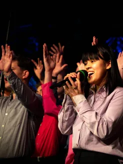 A young singer from the Kölner Jugendchor St. Stephan performs in the foreground with a microphone, wearing a light purple blouse and dark trousers. Behind her, other members of the choir stand in various colorful outfits, including red and green tops, singing along with raised hands and visible enthusiasm. The scene is illuminated by stage lighting, creating an energetic and festive concert atmosphere.