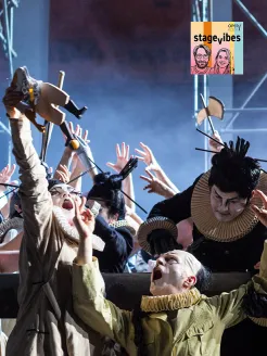 Szenenfoto aus der Oper Turandot: Mehrere Darstellerinnen und Darsteller mit weiß geschminkten Gesichtern und kostümhaften Halskrausen bewegen sich expressiv auf der Bühne. Im Vordergrund hebt eine Person mit weit geöffnetem Mund die Arme, während weitere Ensemblemitglieder im Hintergrund gestisch agieren. Die Szene wirkt energiegeladen und dramatisch. Oben rechts ist das Cover von stage.vibes, dem Podcast der Oper Köln, eingeblendet.