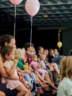 Kinder und Erwachsene sitzen dicht nebeneinander in einer Veranstaltungsreihe und schauen aufmerksam auf eine Darbietung außerhalb des Bildes. Mehrere Kinder sitzen auf dem Schoß ihrer Eltern, während rosa und gelbe Luftballons über den Zuschauenden schweben. Die Atmosphäre wirkt konzentriert und familienfreundlich. Der Veranstaltungsraum ist modern gestaltet, mit einer dunklen Bühne im Hintergrund und einer markanten Rasterdecke.