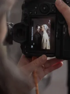 Fotografin Teresa Rothwangl hält eine Kamera in den Händen und betrachtet das gerade aufgenommene Motiv auf dem Kameradisplay. Zu sehen ist ein Porträt zweier Performer*innen, das während des Fotoshootings für das Kampagnenmotiv der Premieren von Der Rosenkavalier der Spielzeit 2026/27 der Oper Köln entstanden ist.