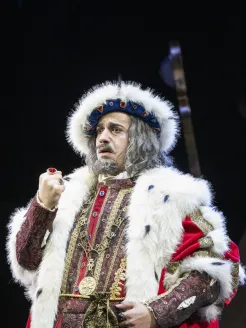 The old King Detlef (Ferhat Baday) raises his ring-adorned fist. He wears a red and gold robe trimmed with white fur, a crown, and a grey wig with a beard. Scene from the children’s opera “DER KÖNIG VERSCHENKT WAS” by Philipp Matthias Kaufmann.