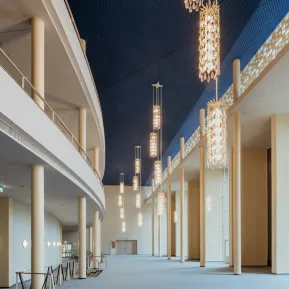 Opernfoyer der Oper Köln mit hohen, hellen Säulen, zwei geschwungenen Galerien und großen Fenstern. Von der dunkelblauen Decke hängen längliche Leuchten mit gläsernen Anhängern in unterschiedlichen Höhen.