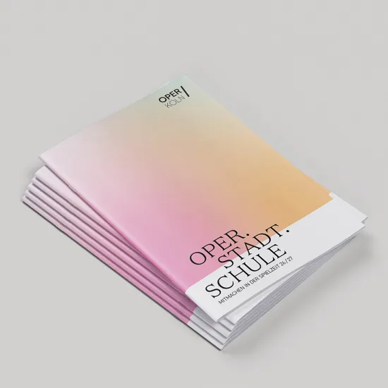 Several copies of an Oper Köln brochure are stacked on a light grey surface. The cover features a soft gradient transitioning from pink to apricot and yellow. The Oper Köln logo appears at the top. In the lower right area, the title “Oper.Stadt.Schule” is displayed, along with the subtitle “Participate in the 2026/27 season.” The brochures are slightly offset, making the individual copies visible.