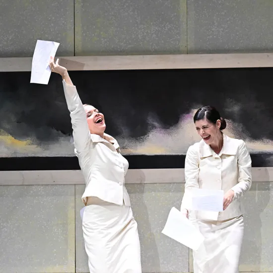Sechs Darsteller*innen aus „Così fan tutte“ auf der Bühne: In der Mitte sitzen zwei Frauen (Fiordiligi und Dorabella) in eleganten, hellen, cremefarbenen, hochgeschlossenen Kleid. Zu ihren Füßen liegen zwei Männer (Ferrando und Guglielmo) in dunklen Tanktops und lässigen Hosen, mit leicht zerzaustem, modernem Erscheinungsbild. Links steht eine Frau (Despina) in einem olivgrünen Hemd und dunklem Rock, rechts ein Mann (Don Alfonso) in einem dunklen Mantel über Hemd und Hose. Die Kostüme verbinden klassische u