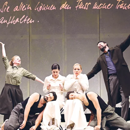 Six performers from Così fan tutte on stage: At the center sit Fiordiligi and Dorabella in elegant, cream-colored, high-necked dresses. At their feet lie Ferrando and Guglielmo, wearing dark tank tops and casual trousers, with a contemporary appearance. To the left stands Despina in an olive-green shirt and dark skirt, while Don Alfonso stands to the right in a dark coat. The costumes combine classical and modern elements.