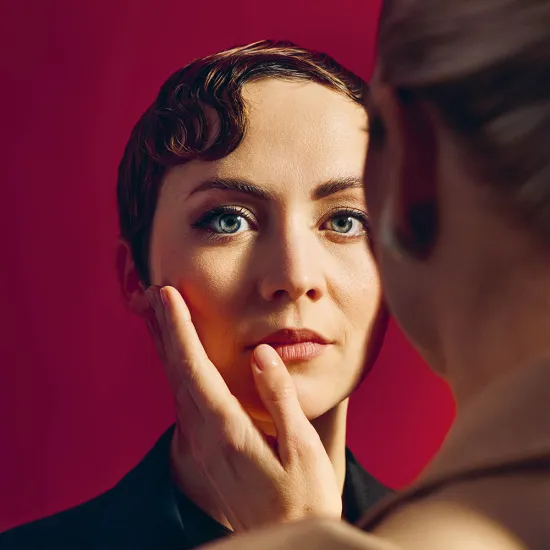 Close-up against a red background with warm, cinematic lighting. At the center stands an androgynous-looking person with short, auburn hair styled in soft finger waves in a dark jacket. Their blue eyes gaze calmly toward the viewer, serious expression appearing thoughtful. In the right foreground, a blonde person with a braid and a light-colored blazer is visible out of focus. Their hand rests gently on the central figure’s cheek. The intimate closeness evokes The Knight of the Rose by Richard Strauss.