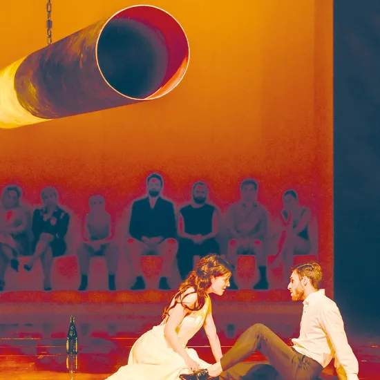 Poppea und Nero in einer Szene aus Die Krönung der Poppea von Claudio Monteverdi: Im Vordergrund kniet Poppea in einem hellen, fließenden Kleid auf dem Bühnenboden und beugt sich intensiv zu Nero hinüber, der ihr gegenüber sitzt, sich nach hinten abstützt und sie aufmerksam ansieht. Die Szene ist in warmes, orangefarbenes Licht getaucht und wirkt sinnlich und spannungsgeladen. Im Hintergrund, leicht erhöht und unscharf dargestellt, sitzt eine Reihe von Figuren nebeneinander und beobachtet das Geschehen. Übe