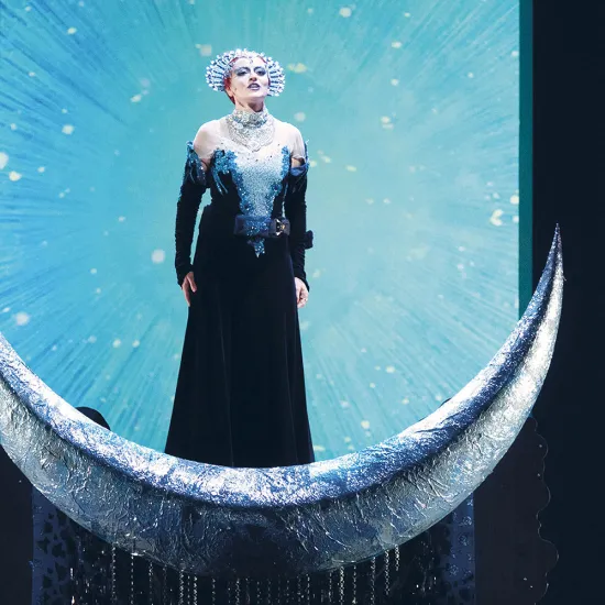 Szene aus Die Zauberflöte von Wolfgang Amadeus Mozart: Die Königin der Nacht steht in einem funkelnden, dunklen Gewand auf einer silbernen Mondsichel, vor einem leuchtenden, sternenartigen Hintergrund; ihre Erscheinung wirkt erhaben und kraftvoll.
