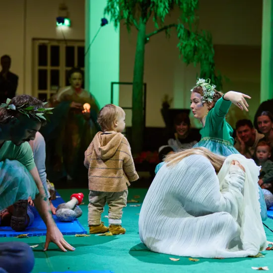 Scene from the opera for very young children Everything That Flies. In a circular performance space, a toddler stands among several performers wearing flowing costumes in shades of green, blue, and white. One performer, wearing a wreath of leaves, gestures expressively toward the child. Parents and children sit around the performance area, experiencing the action at close range. Natural elements, fabrics, and scattered leaves create a poetic and playful atmosphere.