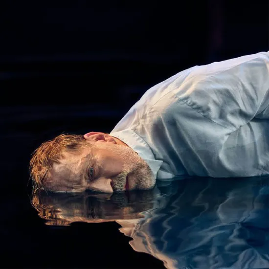 Nahaufnahme eines Mannes mittleren Alters vor schwarzem Hintergrund mit cineastischer Bildwirkung und warmer Lichtstimmung: Mit kurzem, zerzaustem braunem Haar liegt er auf einem wasserbedeckten Boden und blickt zum Betrachter. Er trägt ein weißes Hemd, sein Gesicht und Oberkörper spiegeln sich in der dunklen Wasserfläche. Sein Ausdruck wirkt erschöpft und nach innen gekehrt; die Szene vermittelt eine Atmosphäre von Rückblick und innerer Zerrissenheit. Sie evoziert die Figur des Hoffmann aus Jacques Offenba