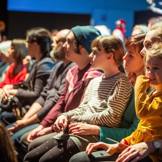 Audience members, including adults and children, sit closely together in a theatre auditorium and watch a performance with focused attention. In the foreground, several children are visible, illuminated by the warm stage light. Their attentive expressions reflect curiosity and engagement. The scene conveys the shared experience of attending a performance at the Kinderoper of Oper Köln.