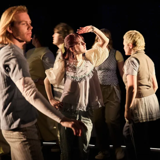 Scene from the performance When I Find Myself in Times of Trouble by the Opera, Song and Scene theatre club: Several young adults move across a dimly lit stage. At the center stands a person with auburn hair, shielding their eyes with one hand as if looking into the distance or searching for direction. Warm stage lighting highlights the central figure, while other participants pass by in different directions. The scene conveys movement, searching, and self-reflection.
