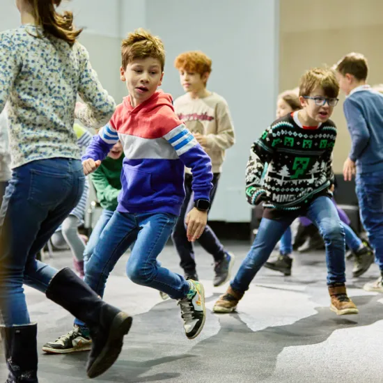 A school class is actively moving through a large indoor space. Several children are running, jumping, and interacting with one another in a playful group exercise. In the foreground, two children can be seen in dynamic motion, while other pupils participate in the background. The scene conveys energy, teamwork, and collaborative learning through movement.