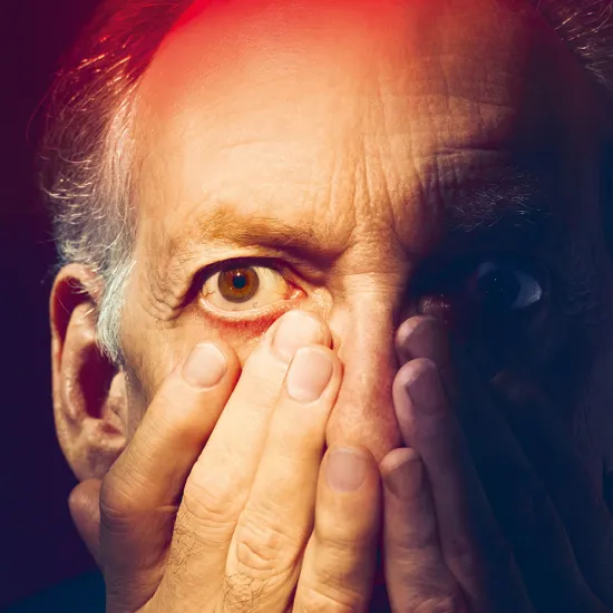 Nahaufnahme eines älteren Mannes mit cineastischer Bildwirkung und warmer Lichtstimmung: Das Gesicht ist frontal zu sehen, der Blick intensiv und weit geöffnet; die Augen wirken erschrocken und angespannt. Die rechte Gesichtshälfte liegt im Schatten. Beide Hände sind vor das Gesicht gelegt, die Finger bedecken Nase, Mund und Wangen – als wolle er sich schützen oder seine Wahrnehmung abschirmen. Die Haut zeigt feine Linien und Falten und glänzt im starken Bühnenlicht. Dramatisches Licht in intensiven Rot- un