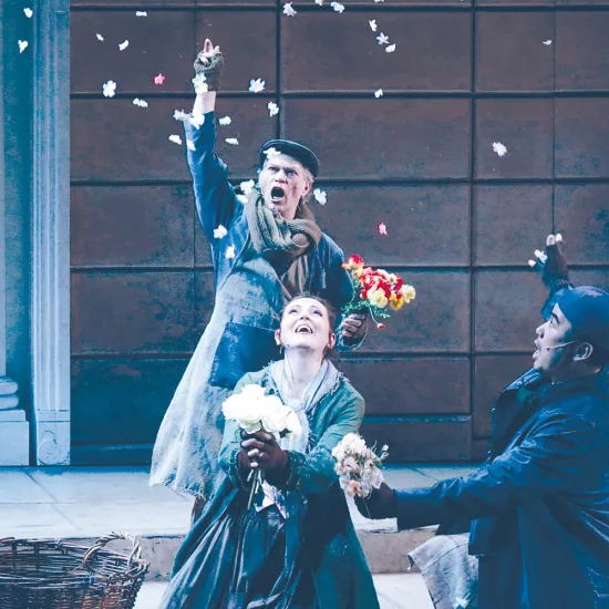 Szene aus My Fair Lady von Frederick Loewe: Drei Darstellende in historisch anmutender Kleidung befinden sich auf einer Straßenkulisse vor einer Hausfassade mit Säulenportal (Covent-Garden-Blumenmarkt): Ein älterer Mann im Hintergrund, gekleidet in Mantel, Schal und Mütze, wirft Blumen in die Luft. Im Vordergrund kniet eine junge Frau, Eliza Doolittle, in einem langen Kleid und hält einen Strauß weißer Rosen, während sie freudig nach oben blickt. Neben ihr kniet ein junger Mann in dunklem Mantel und Schirmm
