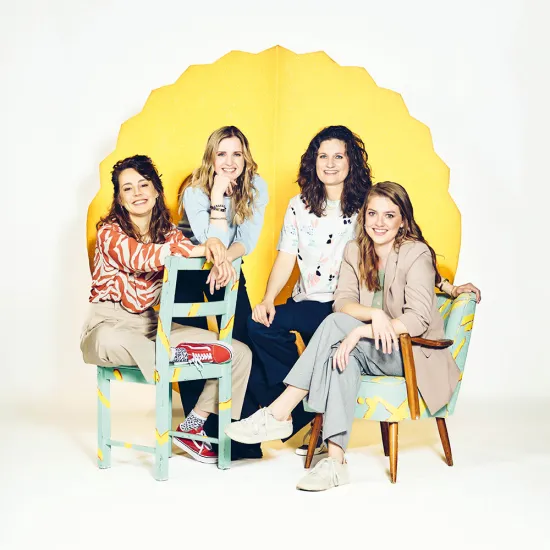 Group portrait of the Oper.Stadt.Schule music theatre education team at Oper Köln. Four women—Nadine Syskowski, Katja Fischer, Stephanie Sonnenschein (Head of Department), and Anna Mangold—are arranged in a bright studio setting against a decorative, glowing yellow backdrop. Two are seated on brightly designed chairs, while the other two stand or lean against them. All are smiling at the camera. Their relaxed poses and the colourful elements create an open, creative, and welcoming impression.