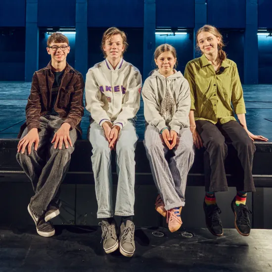 Four members of the Operators youth group of Kinderoper Köln sit side by side on the edge of the stage, looking directly into the camera. In the background, the stage of the new children's opera house at Offenbachplatz can be seen. The teenagers are dressed casually and smile at the camera. The stage and its blue architectural backdrop frame the group portrait.