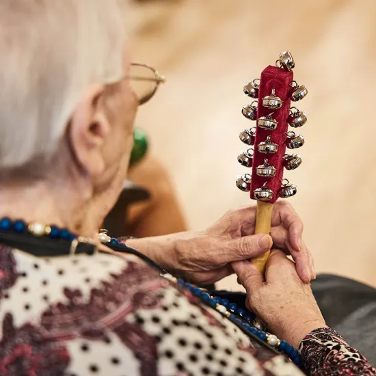 An elderly woman sits in an Oper.Stadt.Schule event and holds a handheld instrument with numerous small bells. The image is taken from behind and focuses on her hands and the instrument. Her calm, concentrated posture conveys active music-making, participation, and the joy of experiencing music together.