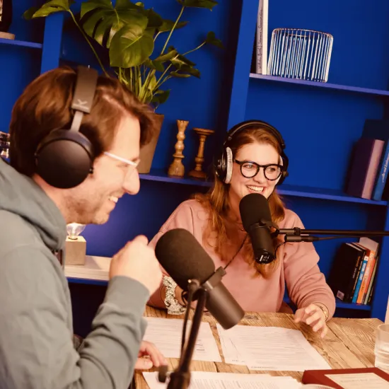 Sebastian Sonntag and Anna Mangold sit at a wooden table in a podcast studio, speaking into microphones. Both are wearing headphones and laughing. Behind them are blue shelves filled with books, plants, and decorative objects. Notes for the recording of the StageVibes podcast are spread out on the table in front of them.