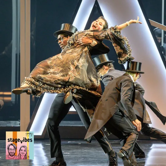 A scene from Rossini’s La Cenerentola: A singer in an elaborate gown is lifted and carried across the stage by performers in dark tailcoats and top hats. Her expressive pose and the dynamic movement create a sense of theatrical energy and exuberance. The StageVibes podcast visual is visible in the lower left corner of the image.