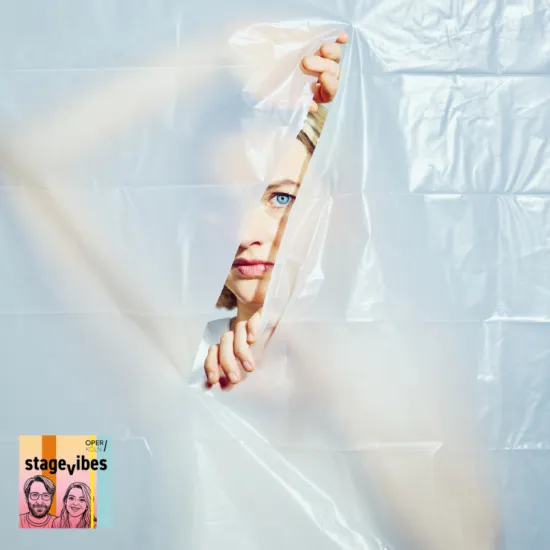 Image from Mozart’s Le Nozze di Figaro: Behind a semi-transparent plastic sheet, a person peers directly at the viewer through a tear in the material. Part of the face, one hand, and a striking blue eye are visible. The composition creates an atmosphere of curiosity, secrecy, and hidden observation. The StageVibes visual appears in the lower left corner of the image.