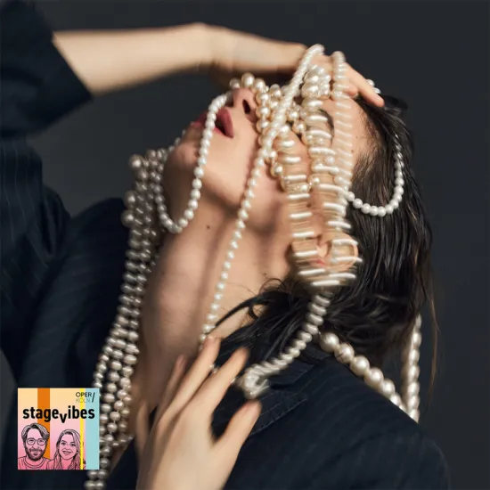Image from Puccini’s Manon Lescaut: A person poses dramatically against a dark background while strands of pearls cascade across their face and shoulders. The expressive gesture and the luxurious jewellery create an atmosphere of glamour, desire, and emotional intensity. The StageVibes visual appears in the lower left corner of the image.