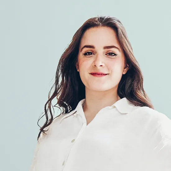 Portrait of the American mezzo-soprano Anna Kelly. She is looking directly into the camera with a slight smile and is wearing a white blouse. Her long dark hair falls loosely over her shoulders against a plain, light-coloured background.