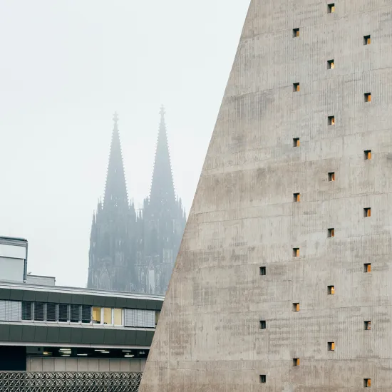 View of the Cologne Opera at Offenbachplatz with its distinctive concrete façade and small square windows. In the background, the twin spires of Cologne Cathedral rise through a light mist. The image highlights the contrast between modern architecture and the city's historic skyline.