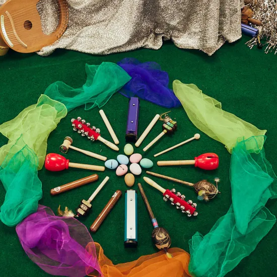 Auf einem grünen Teppich liegen verschiedene Musikinstrumente und Klangobjekte kreisförmig um bunte Eier angeordnet. Trommeln, Rasseln, Klanghölzer, Glöckchen und weitere Instrumente werden von farbigen Tüchern umrahmt. Die Aufnahme zeigt Materialien für ein musikpädagogisches Angebot der Oper.Stadt.Schule.