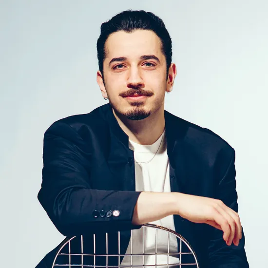 Portrait of bass Ferhat Baday against a light grey studio background. He is seated behind the backrest of a metal chair, resting both arms casually on it while looking directly into the camera with a subtle smile. He is wearing a dark blazer over a white T-shirt. His dark hair is slicked back, and he has a moustache, a goatee, and small earrings. His relaxed posture conveys calmness and confidence.