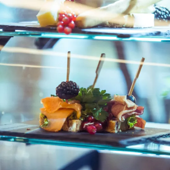 Close-up of a display featuring small open sandwiches topped with smoked salmon, ham, herbs, berries and cocktail picks. The food is presented on dark serving platters inside a glass display case, conveying the high quality of the culinary offering.