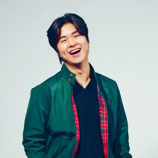 Portrait of tenor Geonmuk Lim against a light grey studio background. He is standing with a slight sideways lean, laughing with his mouth open and looking towards the camera. He is wearing a dark green jacket with a red checked lining over a black top and dark trousers. His medium-length black hair is casually styled. His relaxed posture and warm smile convey openness and joy.
